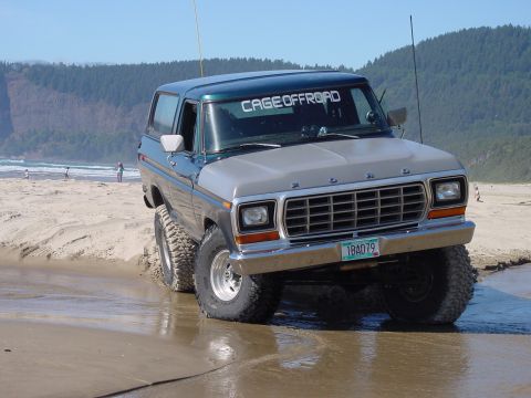 1979 Ford Bronco - A two-tone Ford Bronco navigates a wet, sandy beach with a wooded cliff in the background.