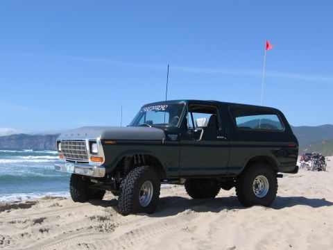 1979 Ford Bronco - A lifted 1979 Ford Bronco in dark green and grey is parked on a sandy beach with the ocean in the background and a red flag flying.