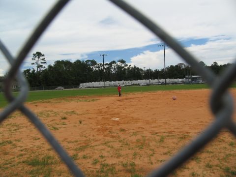 1995 Ford C-350 (Centurion Conversions) - A baseball field is viewed through a chain-link fence, with trees and parked vehicles in the background.
