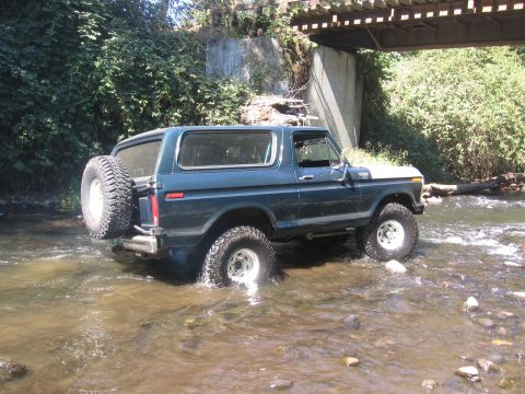 1979 Ford Bronco - A blue 1979 Ford Bronco navigates a shallow stream beneath a wooden bridge.