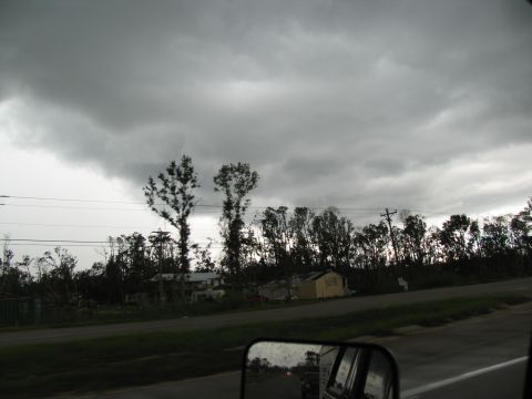 1995 Ford C-350 (Centurion Conversions) - A reflection in a side mirror shows a road and other vehicles under a dark, stormy sky, with damaged trees and buildings visible in the bac…