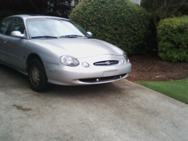 Silver sedan parked on concrete driveway, front view. Bush and trees in background