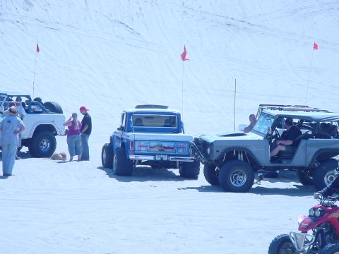 1979 Ford Bronco - People and their off-road vehicles gather on a sand dune under a bright sky.