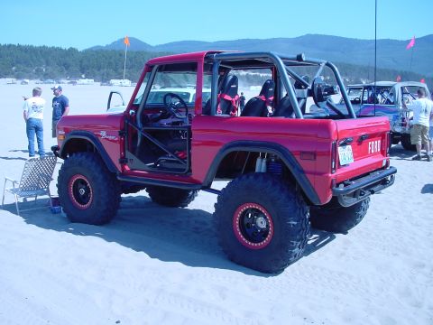1979 Ford Bronco - A red 1979 Ford Bronco modified with a roll cage and oversized tires is parked on a sandy beach, with another vehicle visible in the backgr…