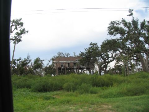 1995 Ford C-350 (Centurion Conversions) - A weathered house stands elevated on stilts amidst lush greenery and trees, suggesting a rural or coastal setting.