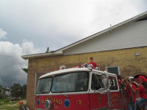 1995 Ford C-350 (Centurion Conversions) - A red fire truck with children on its roof and sides is parked in front of a yellow brick building.