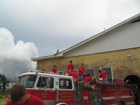 1995 Ford C-350 (Centurion Conversions) - A fire truck with several children on its roof stands in front of a yellow brick building under a cloudy sky.