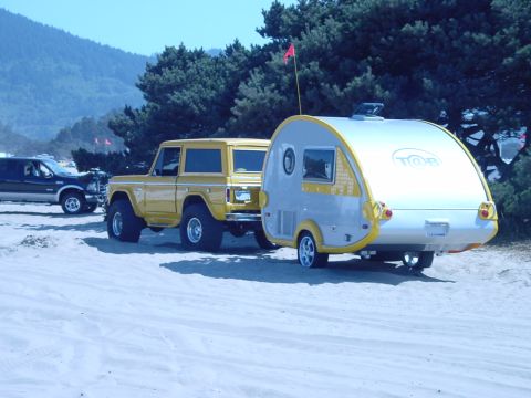 1979 Ford Bronco - A yellow 1978 Ford Bronco tows a silver teardrop trailer on a sandy beach with trees and hills in the background.