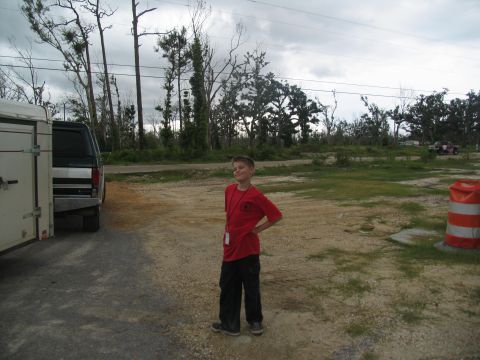 1995 Ford C-350 (Centurion Conversions) - A boy stands next to a silver truck and trailer in a lot with damaged trees in the background.