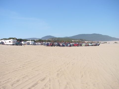 1979 Ford Bronco - A large gathering of vehicles and RVs are parked on a sandy terrain with hills in the background.