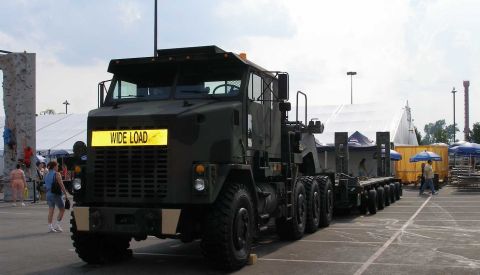 1998 Ford Crown Victoria - A dark olive-green military heavy-haul truck tractor (Oshkosh M1070-style) with a 'WIDE LOAD' sign on the grille, coupled to a multi-axle l…