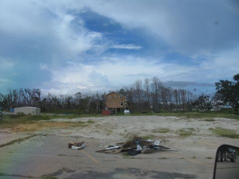 1995 Ford C-350 (Centurion Conversions) - A house elevated on stilts stands amidst debris and damaged trees following a disaster.