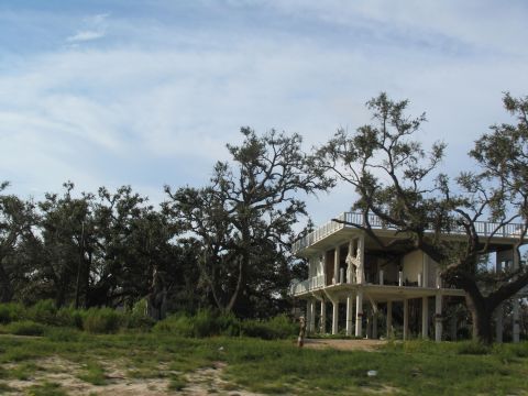 1995 Ford C-350 (Centurion Conversions) - A damaged house on stilts stands amidst trees, showing signs of destruction and neglect.