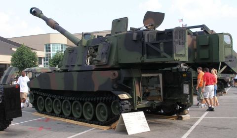 1998 Ford Crown Victoria - Camouflage-painted tracked self-propelled howitzer in the style of an M109, on static display in a parking lot with its long gun barrel rai…