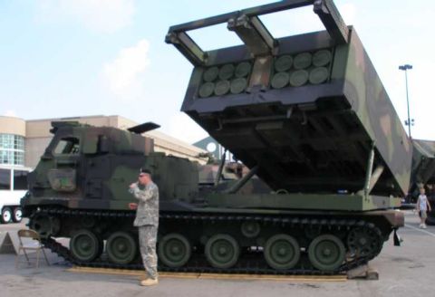 1998 Ford Crown Victoria - A woodland-camouflaged tracked military rocket launcher (M270 MLRS-style) displayed with its rocket-pod launcher assembly raised, a soldier…