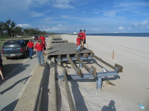 1995 Ford C-350 (Centurion Conversions) - A group of people stand on and near a damaged boardwalk on a beach, with a blue SUV parked nearby.