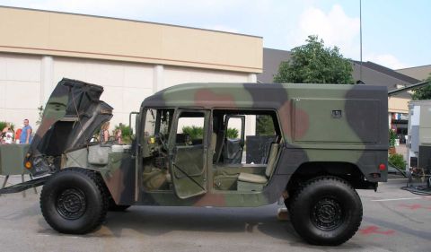 1998 Ford Crown Victoria - Driver-side profile of a military HMMWV (Humvee) in woodland camouflage displayed in a parking lot, with its clamshell hood tilted fully fo…