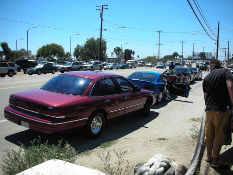 1998 Ford Crown Victoria - maroon Ford sedan parked at the curb beside a blue car in a crowded coastal parking lot, with a person standing at right.