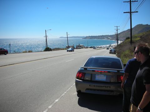1998 Ford Crown Victoria - tan Mustang Cobra parked along a coastal highway with two men standing beside it and the ocean and hills in the background.