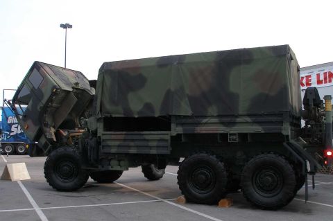1998 Ford Crown Victoria - Side view of a camouflage-painted military 6x6 tactical cargo truck displayed in a parking lot with its cab tilted fully forward to expose…