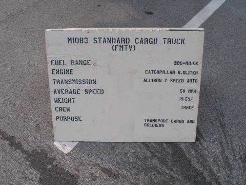 1998 Ford Crown Victoria - Stenciled white display placard propped on pavement listing specifications for the M1083 Standard Cargo Truck (FMTV): 300+ mile fuel range,…