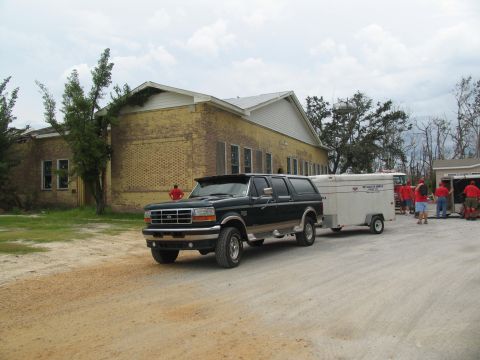 1995 Ford C-350 (Centurion Conversions) - A Ford SUV tows a trailer near a damaged yellow building, with several people in red shirts gathered around.