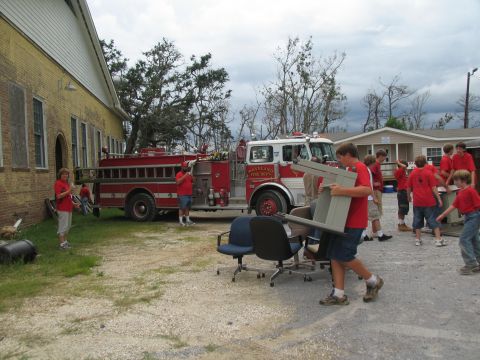 1995 Ford C-350 (Centurion Conversions) - A red fire truck is parked next to a building while people carry furniture, suggesting disaster relief efforts.