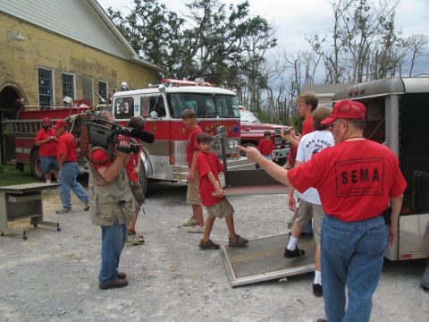 1995 Ford C-350 (Centurion Conversions) - A group of people, some wearing red shirts, are gathered around a fire truck and a trailer.
