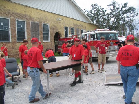 1995 Ford C-350 (Centurion Conversions) - Volunteers in red shirts are seen moving furniture outside a building, with a fire truck in the background.