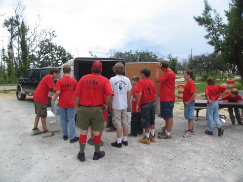 1995 Ford C-350 (Centurion Conversions) - An extended cab pickup truck is parked next to a trailer while a group of people in red shirts unload supplies.