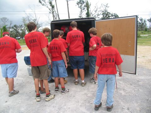 1995 Ford C-350 (Centurion Conversions) - People in red shirts are gathered around an open trailer, appearing to load or unload items.
