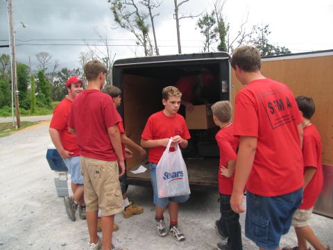 1995 Ford C-350 (Centurion Conversions) - A group of boys are seen helping to unload items from the back of a trailer.