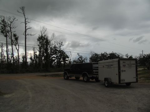 1995 Ford C-350 (Centurion Conversions) - A dark SUV is parked on a dirt road, towing a white cargo trailer under a cloudy sky.