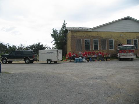 1995 Ford C-350 (Centurion Conversions) - A dark green SUV tows a trailer, while a fire truck and a group of people are gathered outside a building, possibly for a community event o…