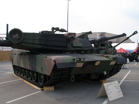 1998 Ford Crown Victoria - M1 Abrams main battle tank in woodland camouflage on static display in a parking lot, viewed from the front three-quarter with its main gun…