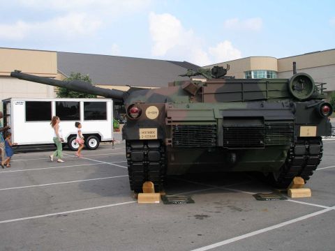1998 Ford Crown Victoria - Front view of a green and brown camouflage M1 Abrams main battle tank displayed on wooden blocks in a parking lot, with its long main gun b…