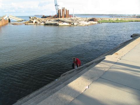 1995 Ford C-350 (Centurion Conversions) - A person kneels by the water near a damaged waterfront with construction equipment in the background.