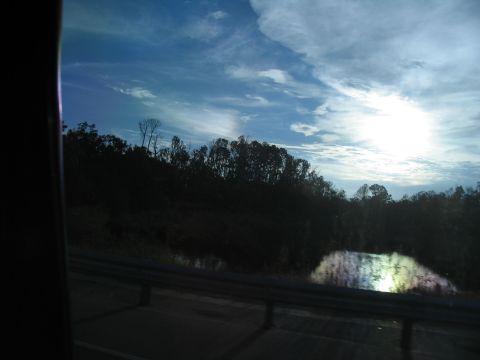 1995 Ford C-350 (Centurion Conversions) - A view from inside a vehicle shows a cloudy sky with sun glare over a dark treeline and reflective water.