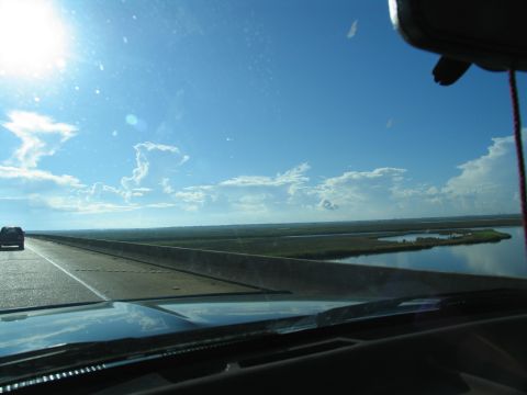 1995 Ford C-350 (Centurion Conversions) - A lone vehicle travels across a bridge, with marshland and water visible to the side under a vast, blue sky.