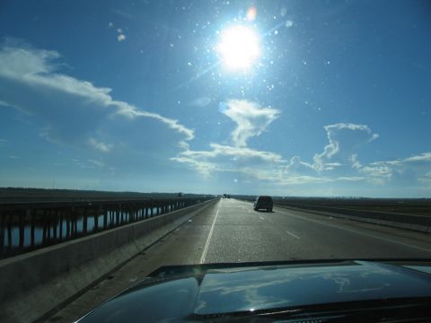 1995 Ford C-350 (Centurion Conversions) - A dark SUV travels down a highway that crosses over a body of water, with a bright sun and scattered clouds in the sky.