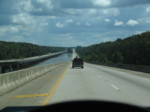 1995 Ford C-350 (Centurion Conversions) - A dark SUV travels down a highway bridge that spans over a body of water, flanked by dense trees under a cloudy sky.