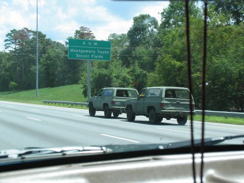 1995 Ford C-350 (Centurion Conversions) - Two olive drab SUVs are seen driving on a highway, passing a green sign for Montgomery Youth Soccer Fields.