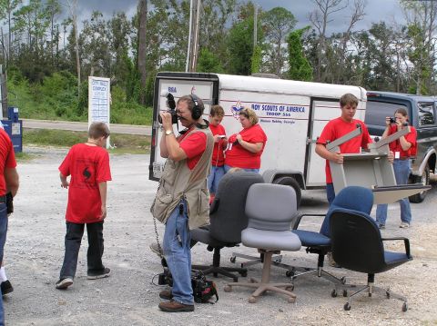 1995 Ford C-350 (Centurion Conversions) - People are gathered around a trailer and a dark van, some filming and others carrying chairs.