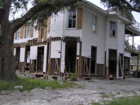 1995 Ford C-350 (Centurion Conversions) - A house shows significant damage to its lower exterior, with siding removed and structural supports visible.