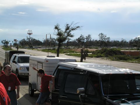 1995 Ford C-350 (Centurion Conversions) - Multiple vehicles and trailers are seen on a road near damaged buildings under a cloudy sky.