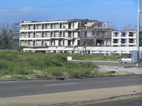 1995 Ford C-350 (Centurion Conversions) - A silver sedan passes by a severely damaged building, its structure exposed to the elements.