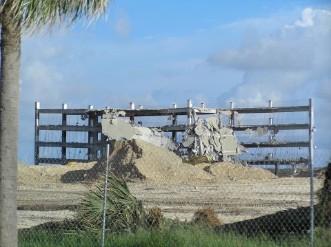 1995 Ford C-350 (Centurion Conversions) - A partially destroyed building stands against a cloudy sky, with debris and sand piles in front.