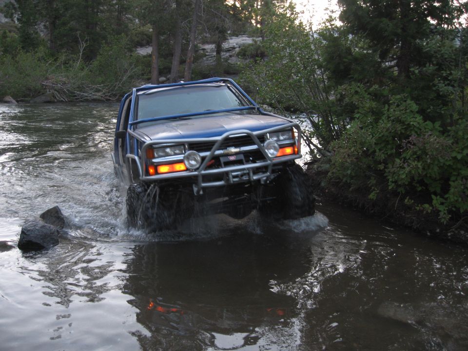 Blue off-road SUV with a roll cage and brush guard driving through a shallow river