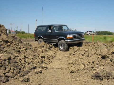 1995 Ford Bronco - A black 1995 Ford Bronco is shown driving through a rough, muddy terrain with piles of dirt on either side.