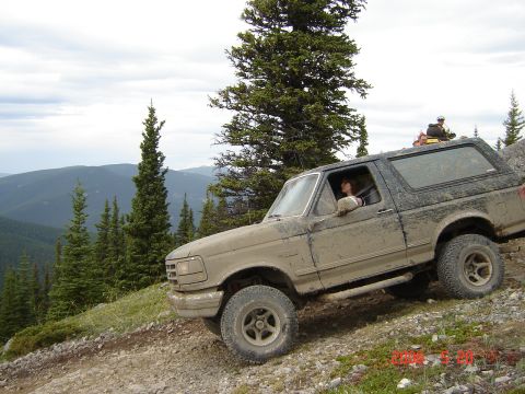 1995 Ford Bronco - A muddy Ford Bronco navigates a steep, rocky off-road path in a mountainous, wooded area.